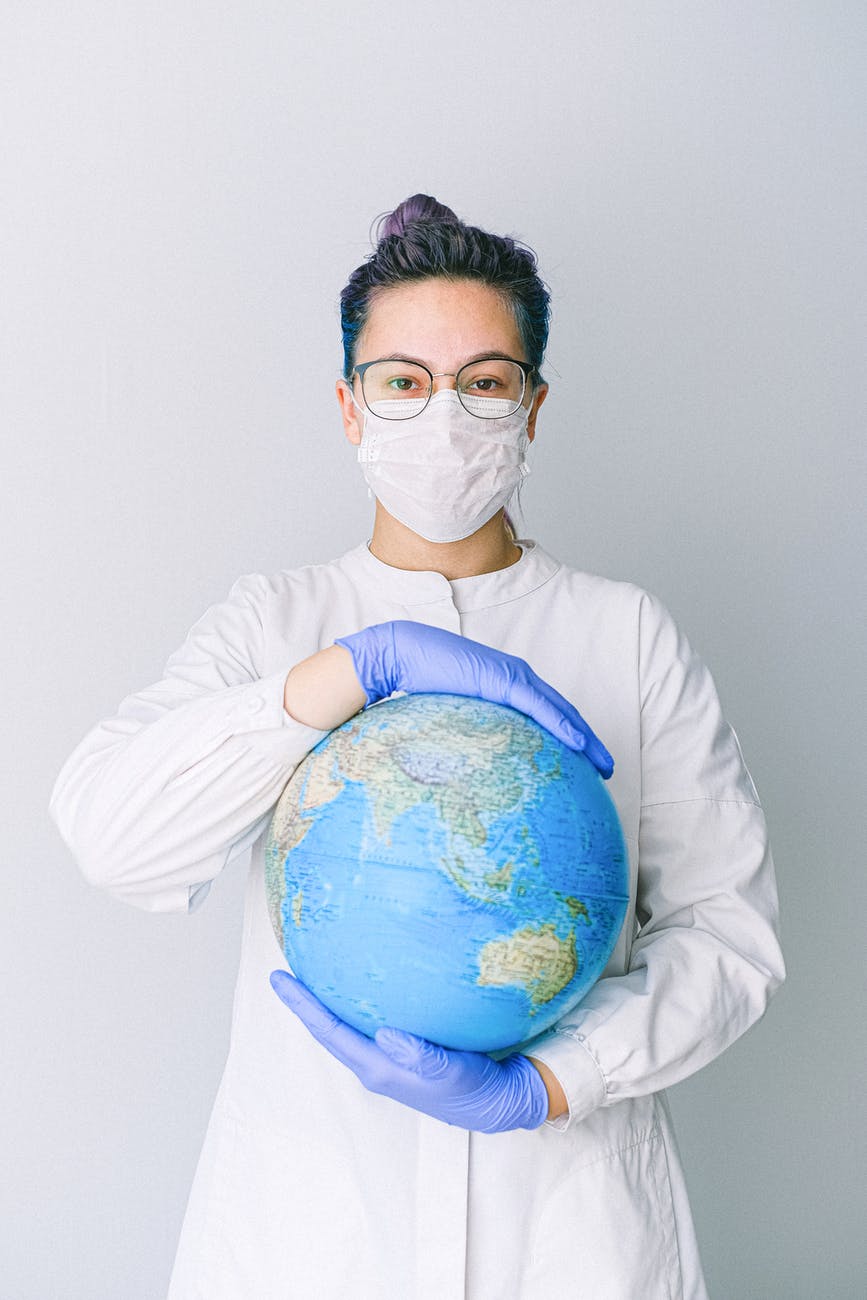 Woman wearing medical mask, gloves, and white coat holds globe in her hands.